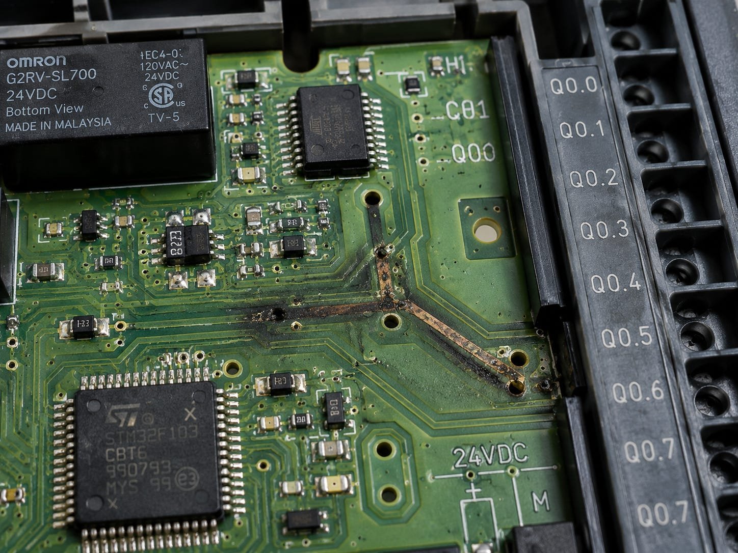 Close-up of a PLC board with a burned, scorched trace running diagonally across it — a fried channel